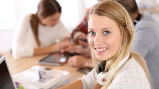 En ung kvinnelig elev sitter på et bord sammen med andre elever. Hun har en PC foran seg.